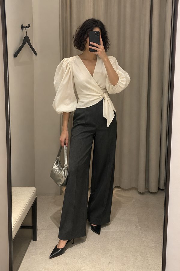 Fitting-room photo of an ivory blouse with sculptural sleeves and a side knot, worn with dark trousers